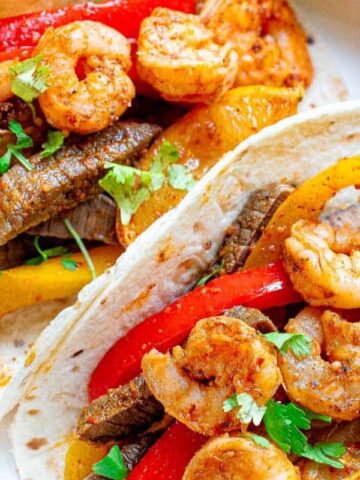 Two tortillas filled with shrimp, beef strips, red and yellow bell peppers, garnished with fresh cilantro on a white plate.