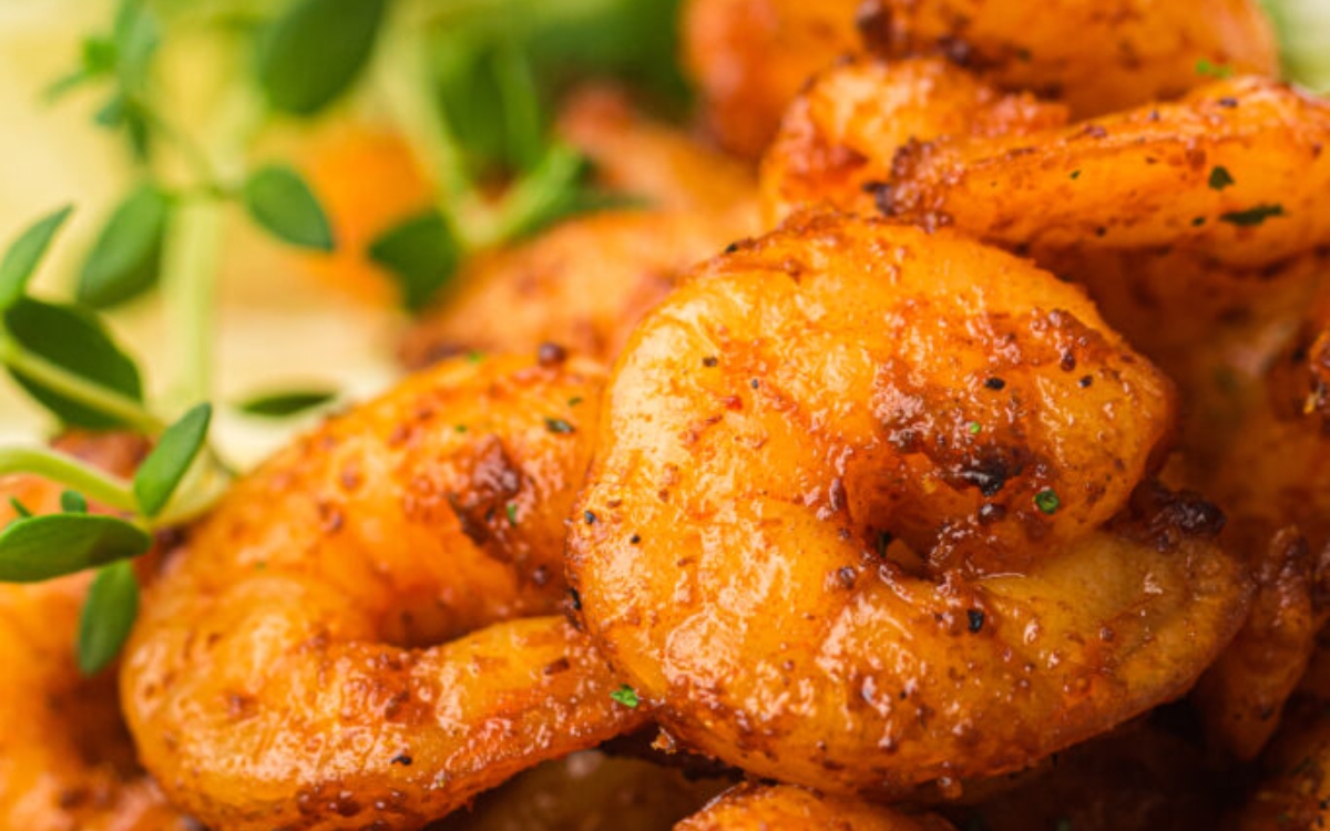 Close-up of cooked, seasoned shrimp garnished with fresh herbs, with blurred lemon slices in the background.