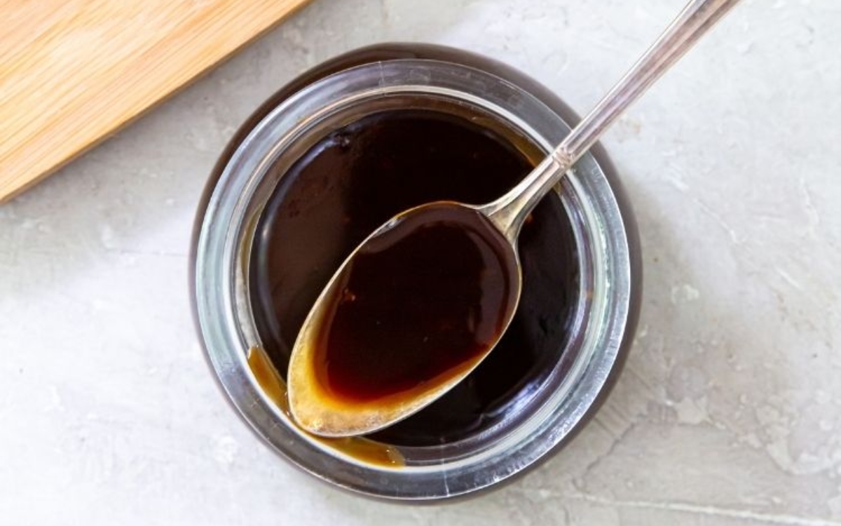 A spoon with dark brown sauce rests on the rim of a glass jar filled with the same sauce, placed on a light-colored surface.