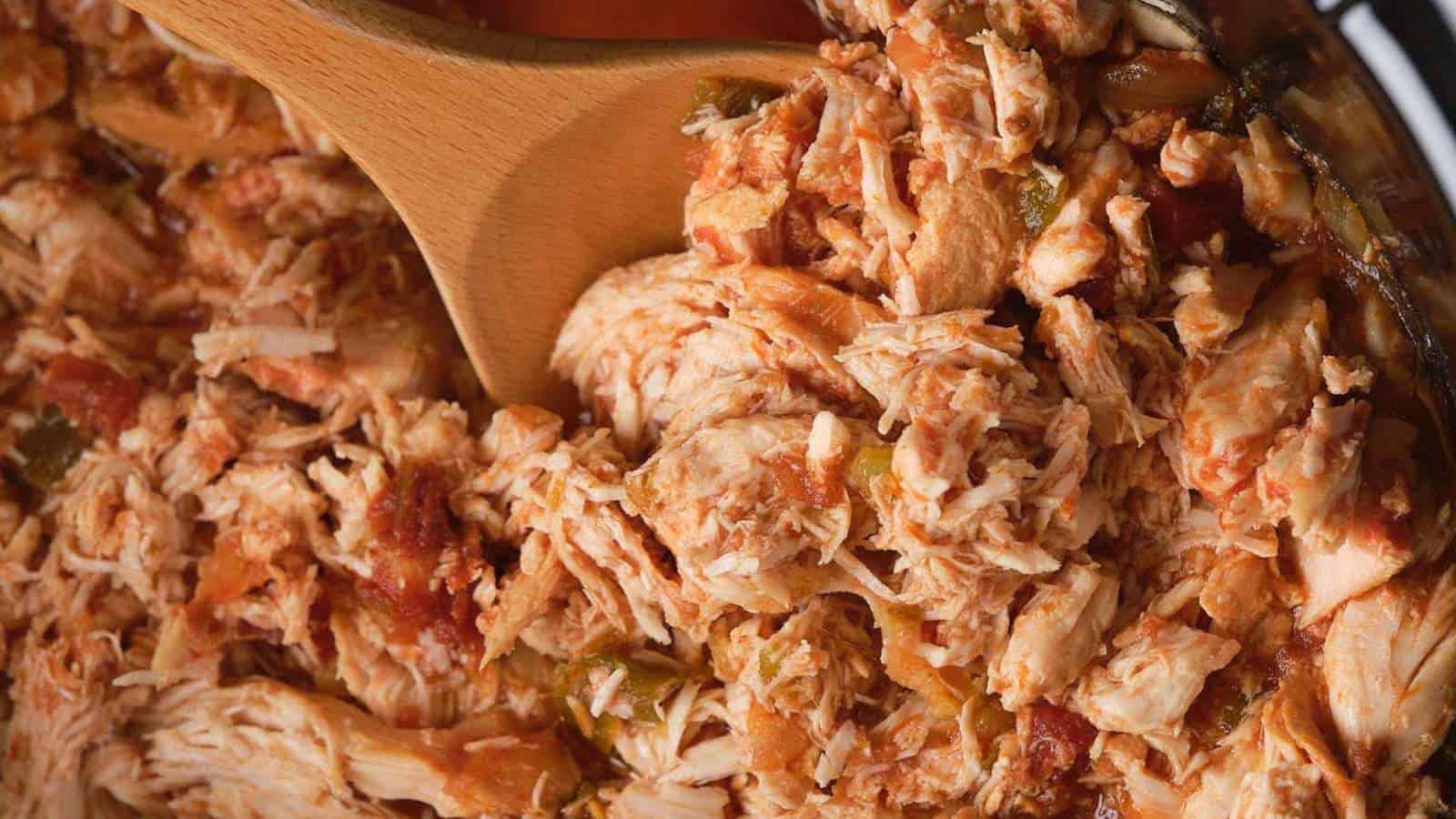 Close-up of shredded chicken with vegetables in a pan, being stirred with a wooden spoon.