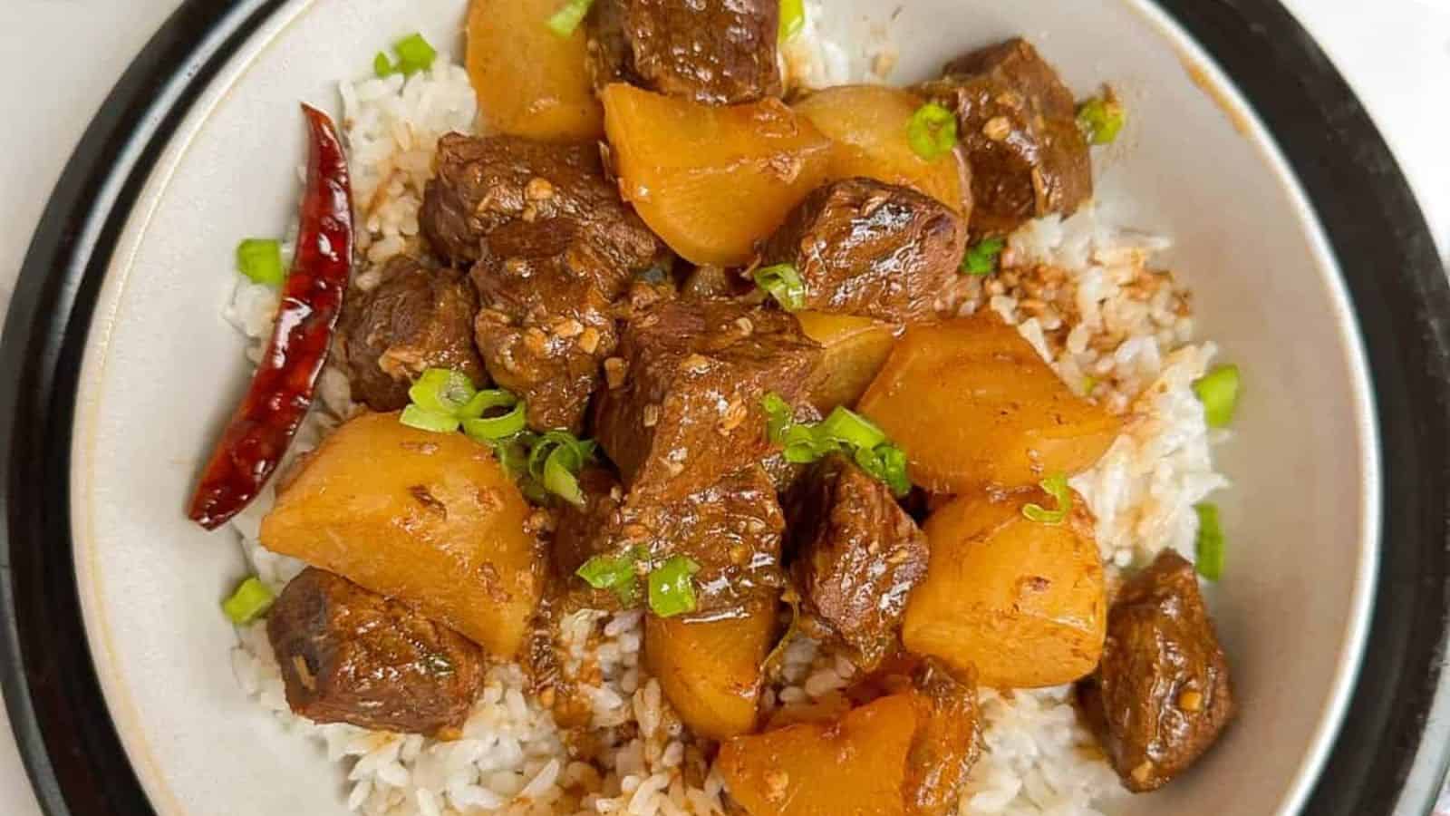 A plate of braised beef and potatoes served on white rice, garnished with chopped green onions and a dried red chili.