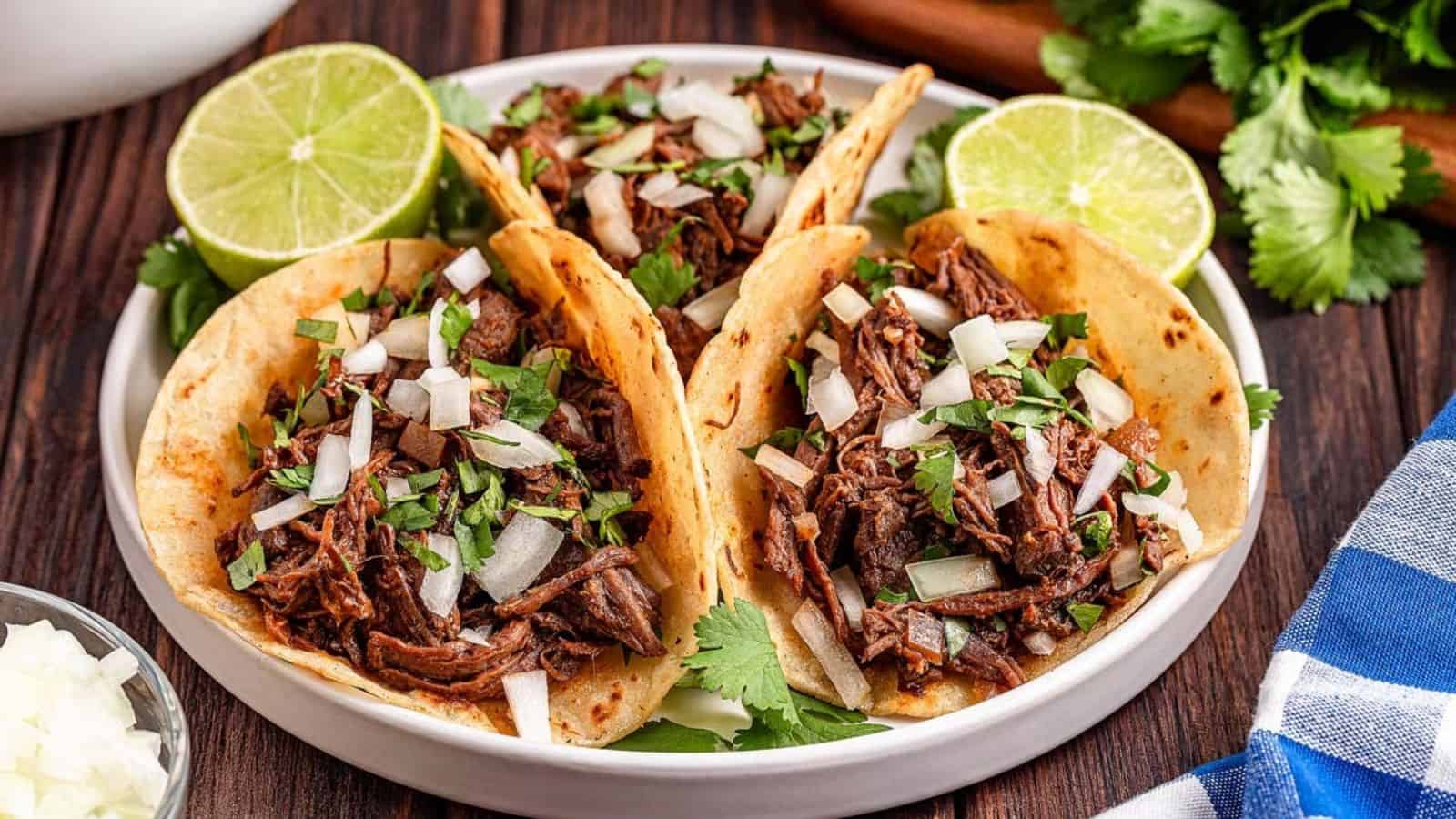 Three beef tacos topped with chopped onions and cilantro served on a white plate with lime halves on the side.