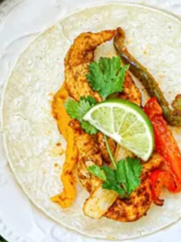 pita wrap top with marinated chicken , pickle, bell pepper and a lime on a plate.