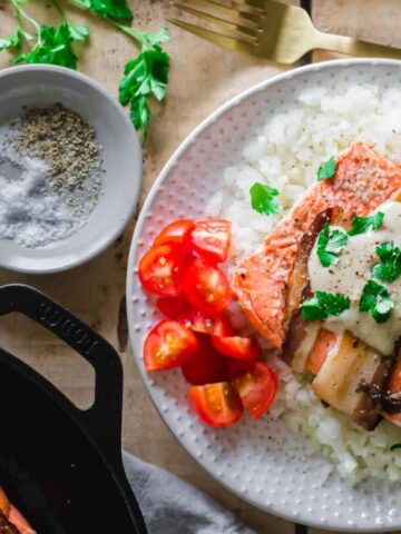 rice top with Bacon Wrapped Salmon and a white sauce garnish with green leaves on a plate.