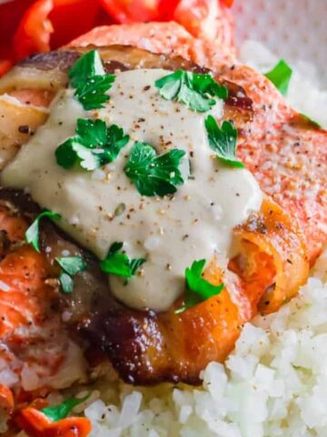 oven baked bacon wrapped salmon recipe topped with tahini sauce and fresh cilantro on a white plate