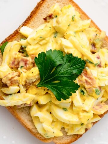 toasted bread top with bacon and egg salad and garnished with a parsley leaf on a plate