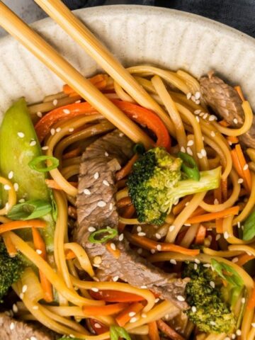 beef, noodles, carrots, broccoli, snow peas, red bell pepper in a bowl with chop stick