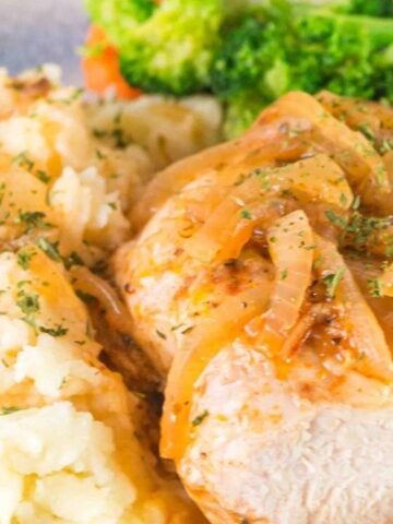 chicken breast topped with onion on a plate with mashed potato and broccoli on the side