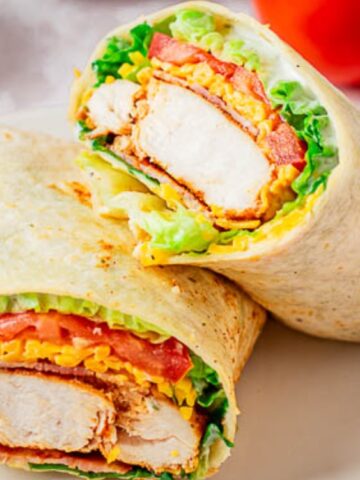 A sliced chicken wrap with lettuce, tomato, and shredded cheese on a white plate, with whole tomatoes in the background.