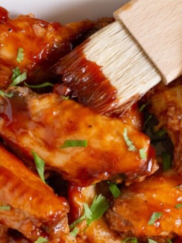 A brush applies barbecue sauce to a plate of chicken wings, garnished with chopped herbs, next to a bowl of extra sauce.