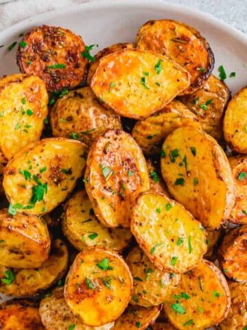 A white plate filled with roasted, halved baby potatoes garnished with chopped parsley, with a spoon and a small bowl of salt nearby.