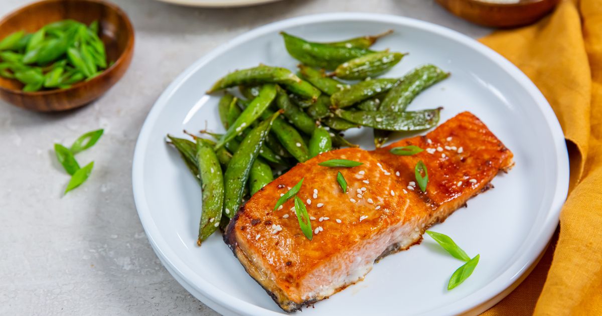 A white plate with a cooked salmon fillet topped with sesame seeds and sliced green onions, served with roasted snap peas.