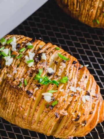 A sliced, seasoned baked potato topped with chopped herbs and sliced garlic on a metal rack inside an air fryer