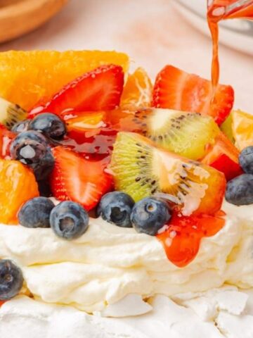 A pavlova dessert topped with whipped cream, strawberries, kiwi, blueberries, orange slices, and a red fruit sauce being poured over the top.