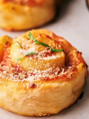 A close-up of a single mini pizza roll topped with grated cheese and green herbs on a light surface.