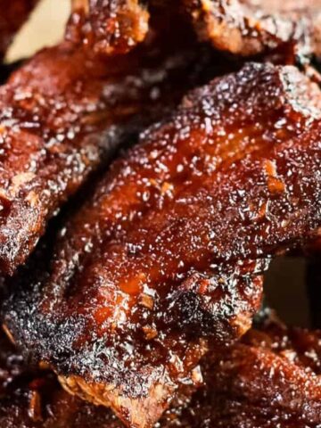 A close-up of several pieces of glazed, cooked barbecue ribs stacked on a wooden surface.