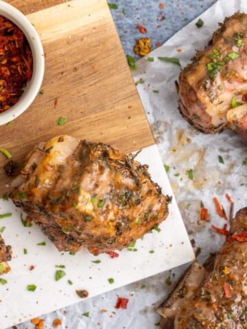 Cooked pork ribs garnished with herbs are arranged on parchment paper and a cutting board, with a small bowl of crushed red pepper flakes nearby.