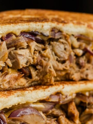 Close-up of a grilled sandwich filled with shredded meat, melted cheese, and sliced red onions on toasted bread.