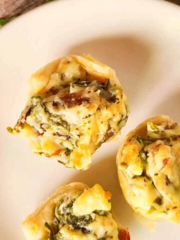 Three mini spinach and cheese pastry bites are arranged on a white plate atop a wooden surface, with some green leafy garnish visible in the background.