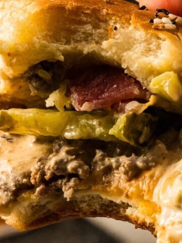 Close-up of a partially eaten cheeseburger with ground beef, melted cheese, pickles, bacon, and sauce inside a soft bun.
