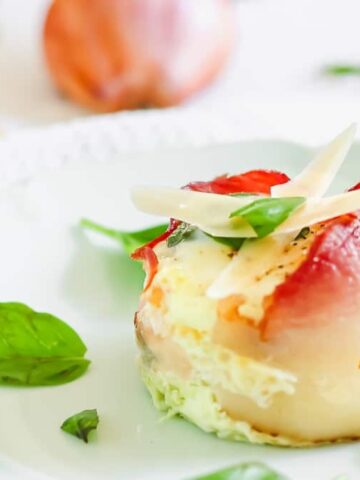 A baked egg cup wrapped in bacon, topped with melted cheese and basil, sits on a white plate garnished with fresh basil leaves.