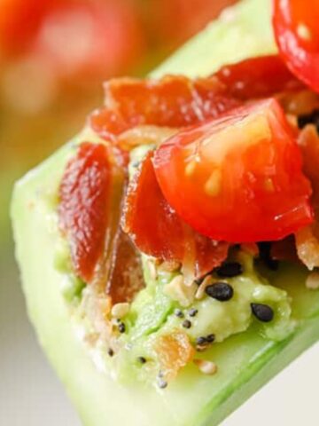 A close-up of a cucumber slice topped with mashed avocado, chopped cherry tomatoes, crumbled bacon, and sprinkled seeds.