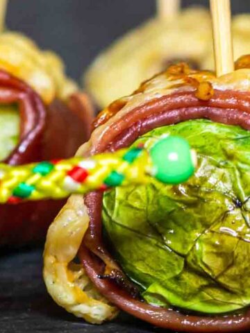 Close-up of Brussels sprouts wrapped in bacon and cheese, secured with skewers, on a dark surface.
