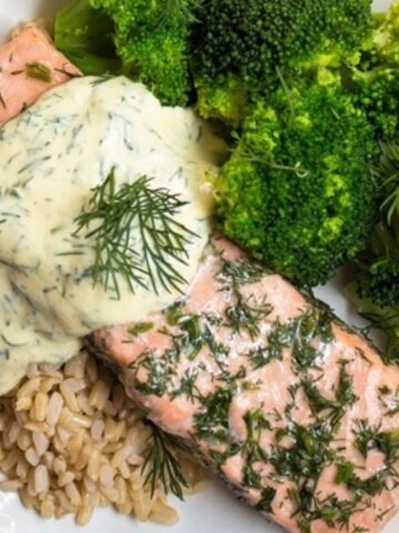 A plate with two pieces of baked salmon topped with creamy dill sauce, served with steamed broccoli and brown rice.