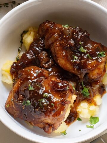 Bowl of glazed chicken thighs served over mashed potatoes, garnished with chopped herbs, on a white surface with a sprig of thyme beside the bowl.