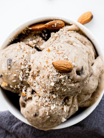 A bowl of creamy ice cream topped with whole almonds and crushed nuts sits on a table beside a spoon and a gray cloth.