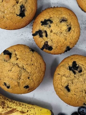 Six blueberry muffins are arranged on a light surface next to a ripe banana and several fresh blueberries.