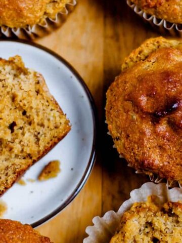 Close-up of several banana muffins on a wooden surface, with one muffin cut in half to show its moist, textured interior.