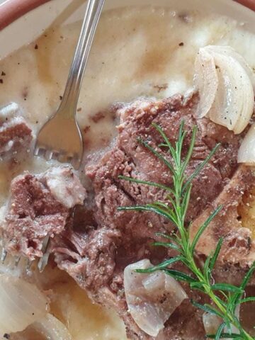 A bowl of creamy soup with a large meat bone, cooked onions, black pepper, and a sprig of fresh rosemary, served with a metal fork.