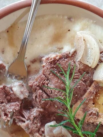 A bowl of creamy soup with pieces of meat, onions, a bone, black pepper, and a sprig of rosemary, with a fork resting on the rim.