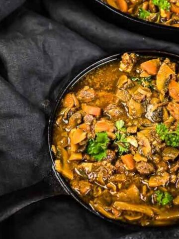 A cast iron skillet filled with beef stew containing chunks of meat, carrots, and mushrooms, garnished with fresh herbs, placed on a dark cloth.