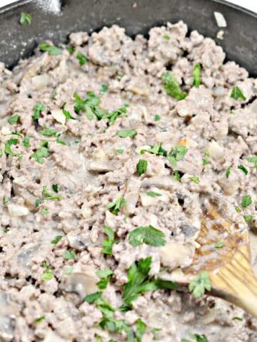 Ground beef and mushrooms cooked in a creamy sauce inside a black skillet, garnished with chopped parsley, with a wooden spoon resting in the mixture.