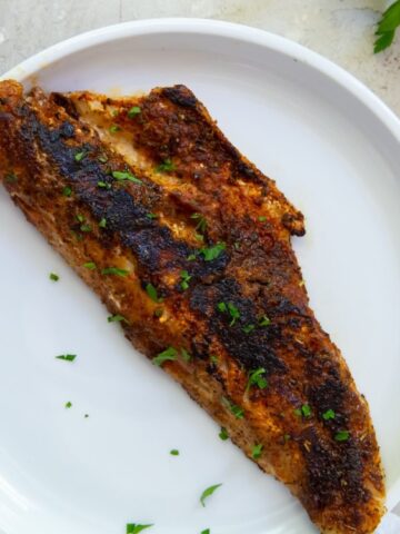 A piece of blackened fish fillet garnished with chopped herbs is served on a white plate.