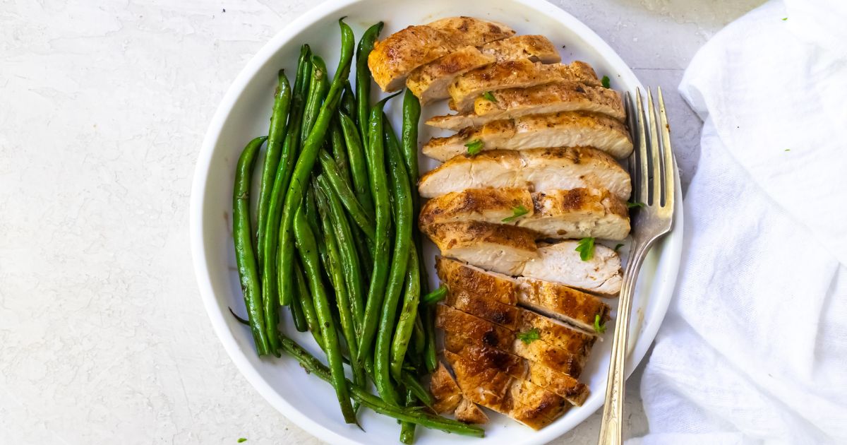 A plate with sliced grilled chicken breast and cooked green beans, served with a fork on a white surface.