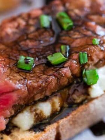 A slice of rare steak on toasted bread with crumbled cheese, topped with chopped chives and a drizzle of dark sauce.