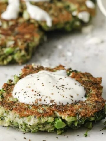 Vegetable fritters topped with sour cream and black pepper, served on a white surface with additional fritters in the background.