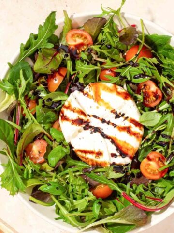 A bowl of mixed greens with cherry tomatoes, a round of grilled cheese in the center, and a drizzle of balsamic glaze, with basil leaves and dressing on the side.
