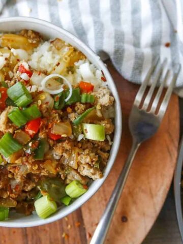 A bowl of rice topped with ground meat, diced red bell peppers, chopped green onions, and cooked onions, placed on a wooden surface next to a fork and a striped napkin.