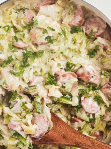 A skillet filled with sliced sausage, cabbage, and creamy sauce, garnished with chopped parsley, with a wooden spoon resting in the dish.