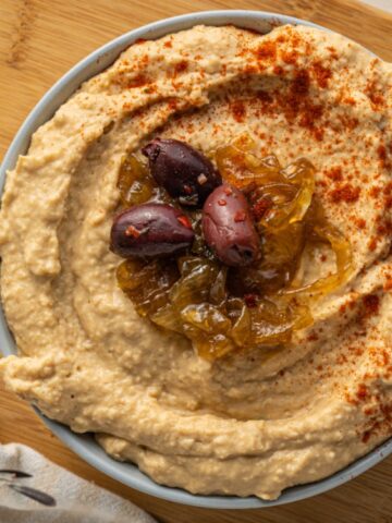 A bowl of hummus topped with caramelized onions, olives, and paprika sits on a wooden cutting board.