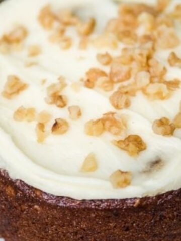 A round carrot cake with white cream cheese frosting and chopped walnuts on top, displayed on a white plate.