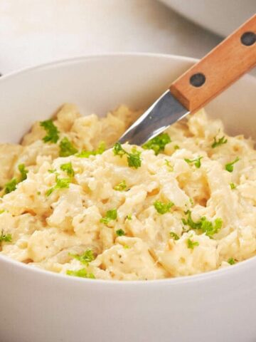 A white bowl filled with creamy, cheesy hashbrown casserole, garnished with chopped parsley, sits on a plate with a wooden-handled spoon resting inside.