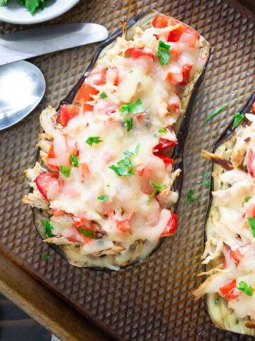 Two baked eggplant halves topped with melted cheese, diced tomatoes, herbs, and shredded chicken on a baking tray beside two spoons and fresh parsley.