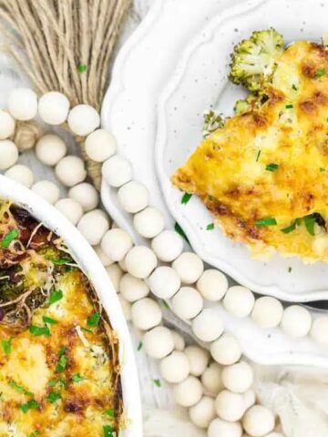 A baked broccoli casserole topped with melted cheese, served on a white plate beside a dish, with decorative wooden beads in the background.