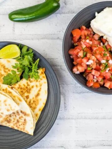 A plate with quesadilla slices, lime, cilantro, and jalapeño slices next to a bowl of pico de gallo and a scoop of sour cream on a white background.