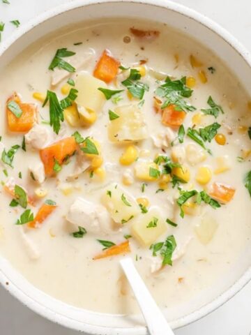 A bowl of creamy soup with chunks of chicken, carrots, corn, potatoes, and chopped parsley on top, served on a white surface.